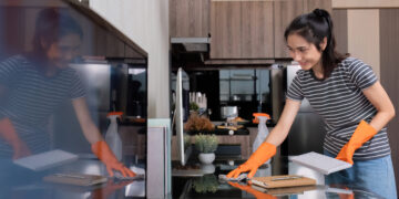 landlord wearing orange gloves cleans a desk in a modern kitchen using a spray bottle and cloth.