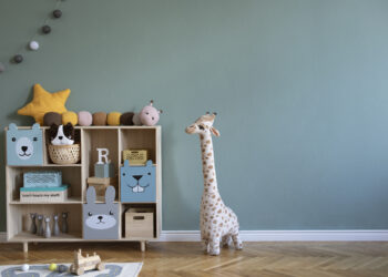 A tidy children's room with a wooden toy shelf, plush animals, a stuffed giraffe, and a pastel green wall.