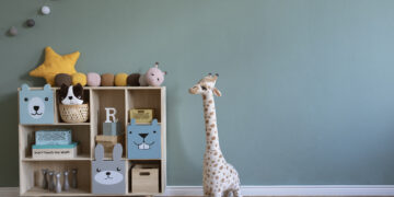 A tidy children's room with a wooden toy shelf, plush animals, a stuffed giraffe, and a pastel green wall.