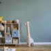 A tidy children's room with a wooden toy shelf, plush animals, a stuffed giraffe, and a pastel green wall.