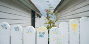 White fence with hand-painted birds, flowers, and sun between two buildings.