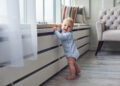 Toddler standing near a low windowsill in a living room, highlighting the need for window safety and furniture securing in child-proofing.