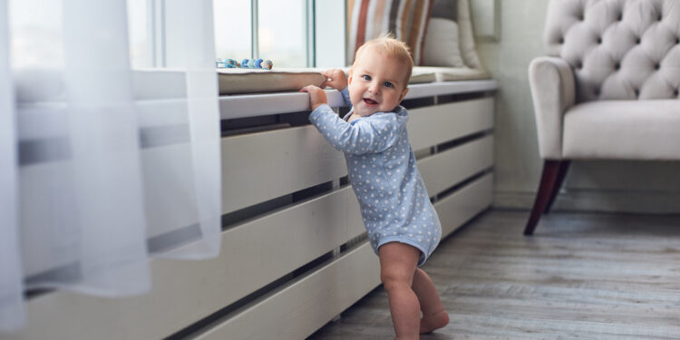 Toddler standing near a low windowsill in a living room, highlighting the need for window safety and furniture securing in child-proofing.