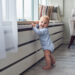 Toddler standing near a low windowsill in a living room, highlighting the need for window safety and furniture securing in child-proofing.