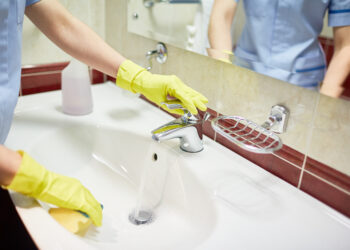 Person wearing gloves cleaning a bathroom sink with running water.
