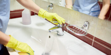 Person wearing gloves cleaning a bathroom sink with running water.