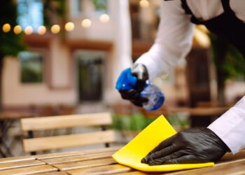 Person wearing black gloves cleaning an outdoor wooden table with a yellow cloth while spraying cleaning solution from a blue bottle.