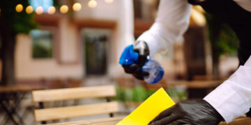 Person wearing black gloves cleaning an outdoor wooden table with a yellow cloth while spraying cleaning solution from a blue bottle.