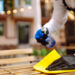 Person wearing black gloves cleaning an outdoor wooden table with a yellow cloth while spraying cleaning solution from a blue bottle.