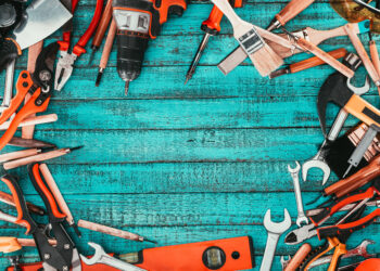 A flat lay of essential DIY tools including a cordless drill, pliers, screwdrivers, paintbrushes, spanners, and a spirit level, arranged on a bright blue wooden surface — ideal for UK homeowners building a basic toolkit.