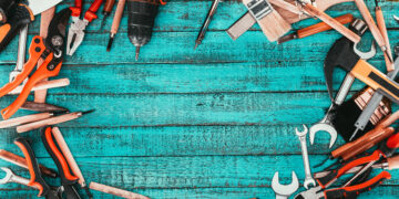 A flat lay of essential DIY tools including a cordless drill, pliers, screwdrivers, paintbrushes, spanners, and a spirit level, arranged on a bright blue wooden surface — ideal for UK homeowners building a basic toolkit.