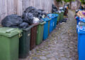 Overflowing outdoor bins and rubbish bags in a residential alley during summer.