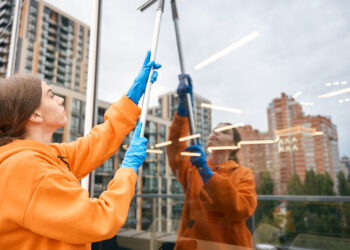 Woman cleaning tall window with squeegee on balcony.