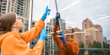 Woman cleaning tall window with squeegee on balcony.