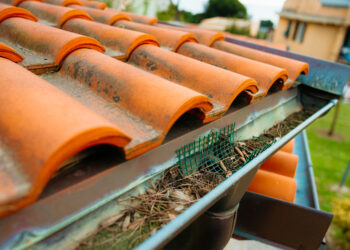A person cleaning fallen leaves from a house gutter in October to prevent blockages and water damage.