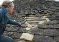 A roofer inspecting and measuring a large gap in a traditional stone-tiled roof, where several loose and missing tiles expose the wooden battens beneath.