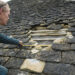 A roofer inspecting and measuring a large gap in a traditional stone-tiled roof, where several loose and missing tiles expose the wooden battens beneath.