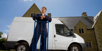 Man in work overalls leaning on a hand truck beside a white removal van outside a residential property in the UK