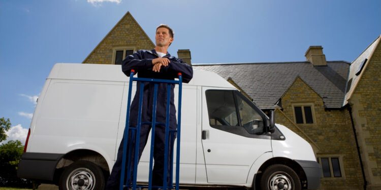 Man in work overalls leaning on a hand truck beside a white removal van outside a residential property in the UK