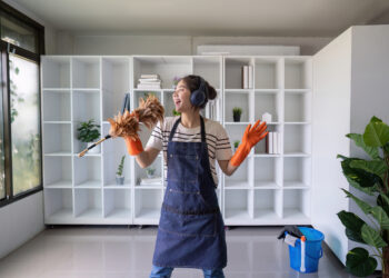 Young woman wearing headphones and cleaning gloves, dancing while holding a feather duster in a bright modern living room with white shelves and a blue cleaning bucket nearby.