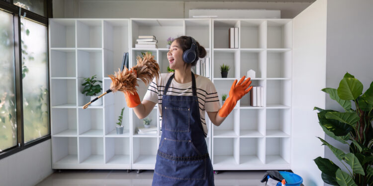 Young woman wearing headphones and cleaning gloves, dancing while holding a feather duster in a bright modern living room with white shelves and a blue cleaning bucket nearby.