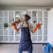 Young woman wearing headphones and cleaning gloves, dancing while holding a feather duster in a bright modern living room with white shelves and a blue cleaning bucket nearby.