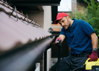 Professional worker cleaning house gutters using a vacuum hose to remove leaves and debris from the roof edge.