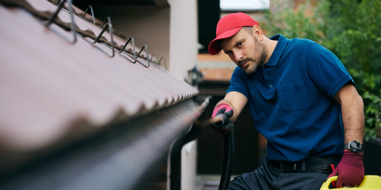 Professional worker cleaning house gutters using a vacuum hose to remove leaves and debris from the roof edge.