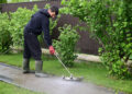 Person using a pressure washer to clean a garden pathway beside a lawn and hedge in a residential outdoor setting.