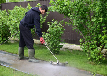 Person using a pressure washer to clean a garden pathway beside a lawn and hedge in a residential outdoor setting.