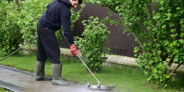 Person using a pressure washer to clean a garden pathway beside a lawn and hedge in a residential outdoor setting.