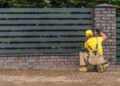 Worker installing horizontal metal fence panels beside a brick pillar in a garden boundary