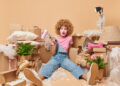 Woman surrounded by moving boxes and packing materials looking overwhelmed while holding a paint roller during house clearance preparation