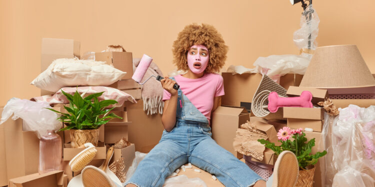 Woman surrounded by moving boxes and packing materials looking overwhelmed while holding a paint roller during house clearance preparation