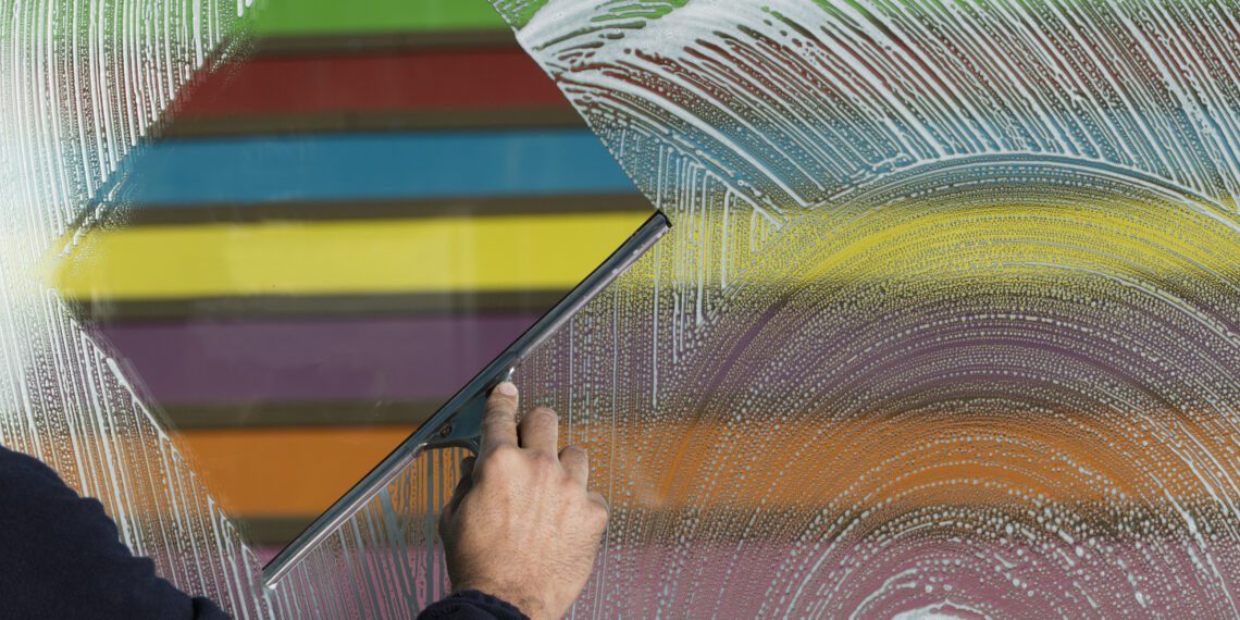 Person using a squeegee to clean a soapy window with colourful horizontal stripes visible through the glass.