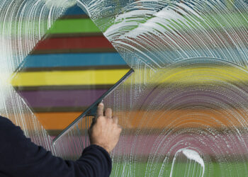 Person using a squeegee to clean a soapy window with colourful horizontal stripes visible through the glass.