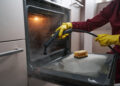 Professional oven cleaner using a steam tool to remove grease from the inside of a kitchen oven.