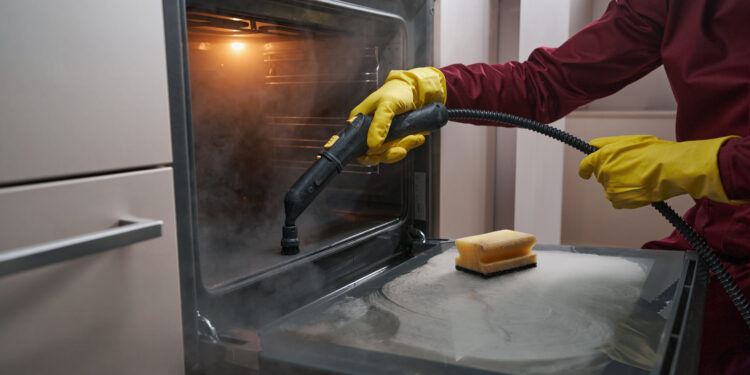 Professional oven cleaner using a steam tool to remove grease from the inside of a kitchen oven.