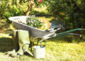 Wheelbarrow filled with flowers and gardening tools beside green rubber boots and a watering can in a sunny garden.