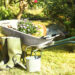 Wheelbarrow filled with flowers and gardening tools beside green rubber boots and a watering can in a sunny garden.
