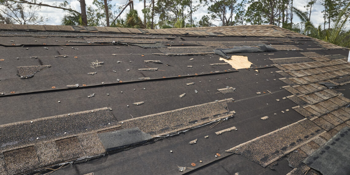 Wind-damaged house roof with missing and torn asphalt shingles exposing underlayment