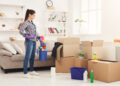 Woman wearing gloves holding a cleaning bucket while standing beside packed cardboard boxes in a bright living room, preparing for end-of-tenancy cleaning.