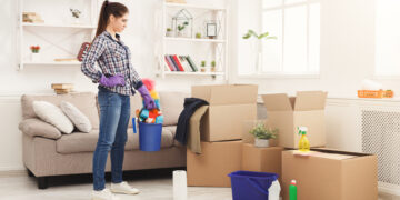 Woman wearing gloves holding a cleaning bucket while standing beside packed cardboard boxes in a bright living room, preparing for end-of-tenancy cleaning.
