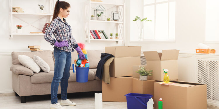 Woman wearing gloves holding a cleaning bucket while standing beside packed cardboard boxes in a bright living room, preparing for end-of-tenancy cleaning.