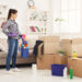 Woman wearing gloves holding a cleaning bucket while standing beside packed cardboard boxes in a bright living room, preparing for end-of-tenancy cleaning.
