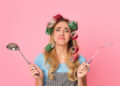 Frustrated woman with hair rollers holding kitchen utensils symbolising common household jobs people put off at home.