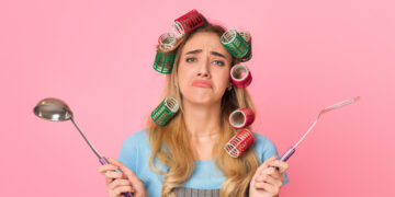 Frustrated woman with hair rollers holding kitchen utensils symbolising common household jobs people put off at home.