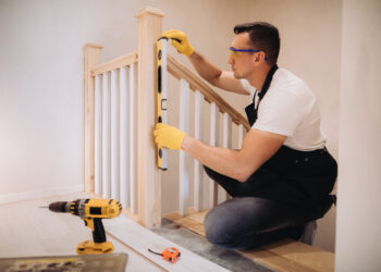 Close-up of a loose wooden stair banister being tightened with a screwdriver, showing slight movement at the wall fixing.