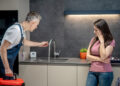 Emergency plumber inspecting leaking kitchen tap while concerned homeowner looks on in modern UK kitchen