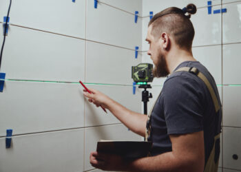 Close-up of a person regrouting bathroom tiles using a grout float, removing old grout and applying fresh sealant between white ceramic tiles.